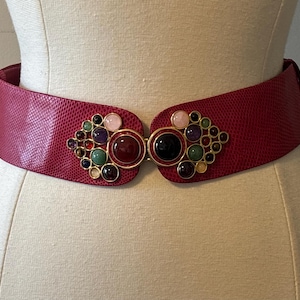 Può includere: Una cintura larga rossa con un motivo testurizzato simile alla pelle di serpente, dotata di una fibbia decorativa in metallo dorato ornata da una serie di pietre cabochon rotonde multicolori nelle tonalità rosa, viola, verde, nero e ambra.