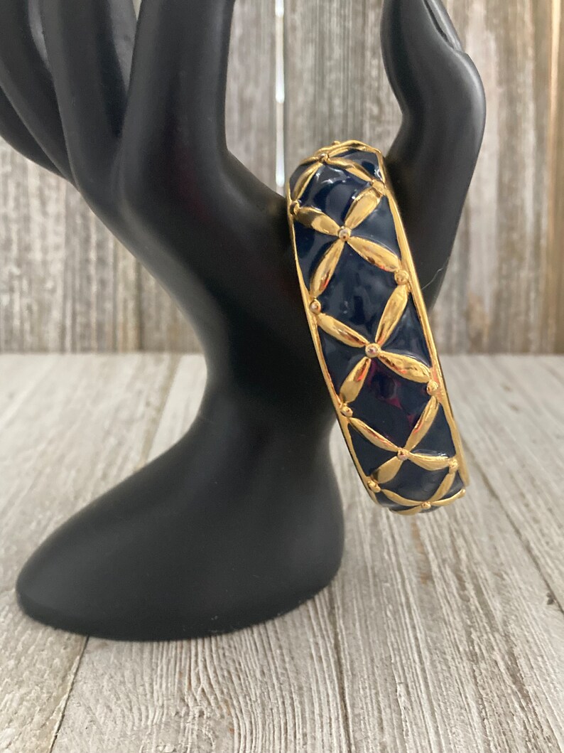 May include: A gold and navy blue bangle bracelet with a geometric pattern. The bracelet is made of metal and has a shiny finish.