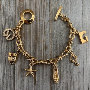 May include: Gold-toned charm bracelet featuring various charms, including a theater mask, a musical note, a ballet slipper, a starfish, and a letter "G". The bracelet has a toggle clasp and a chain link design.