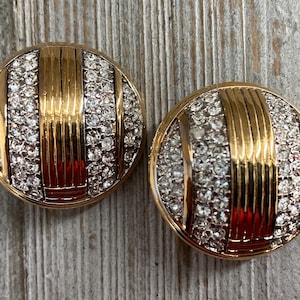 Puede incluir: Un par de pendientes de clip de tono dorado con un diseño circular. Los pendientes están adornados con diamantes de imitación transparentes y rayas doradas.