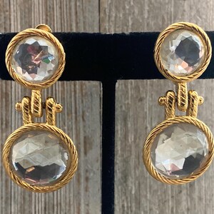 Pode incluir: Um par de brincos de gota em tom dourado com pedras de cristal facetadas transparentes. Os brincos t&ecirc;m um design de corda em torno das pedras.