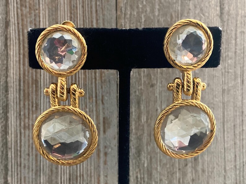 Pode incluir: Um par de brincos de gota em tom dourado com pedras de cristal facetadas transparentes. Os brincos t&ecirc;m um design de corda em torno das pedras.