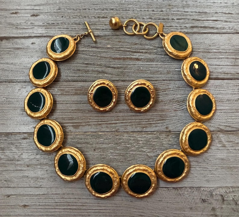 K&ouml;nnte beinhalten: Ein goldfarbenes Halsketten- und Ohrring-Set. Die Halskette hat gro&szlig;e, kreisf&ouml;rmige, dunkelgr&uuml;ne Steine, die in einen strukturierten Goldrahmen gefasst sind. Die Ohrringe haben ein &auml;hnliches Design. Der Schmuck wird auf einer Holzoberfl&auml;che pr&auml;sentiert.
