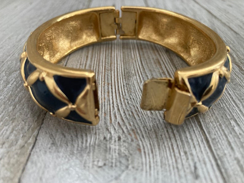 May include: Gold tone hinged bangle bracelet with a blue enamel design. The bracelet has a geometric pattern with a floral motif.