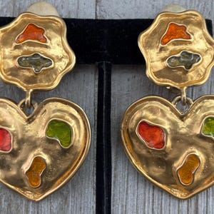 May include: A pair of gold-toned clip-on earrings. Each earring features a flower-shaped top and a heart-shaped bottom. The earrings have colorful enamel accents in red, orange, and green. The earrings are displayed on a black stand.