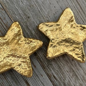 May include: Two gold-colored star-shaped decorative objects with textured surfaces. The stars are placed on a wooden surface. The stars have a metallic finish and are likely made of metal or a similar material.
