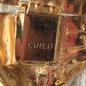 Pu&ograve; includere: Primo piano di un gioiello dorato con la scritta "CINER" stampata su una placca rettangolare. Il gioiello presenta un design ornato e strutturato con dettagli intricati e una finitura lucida.