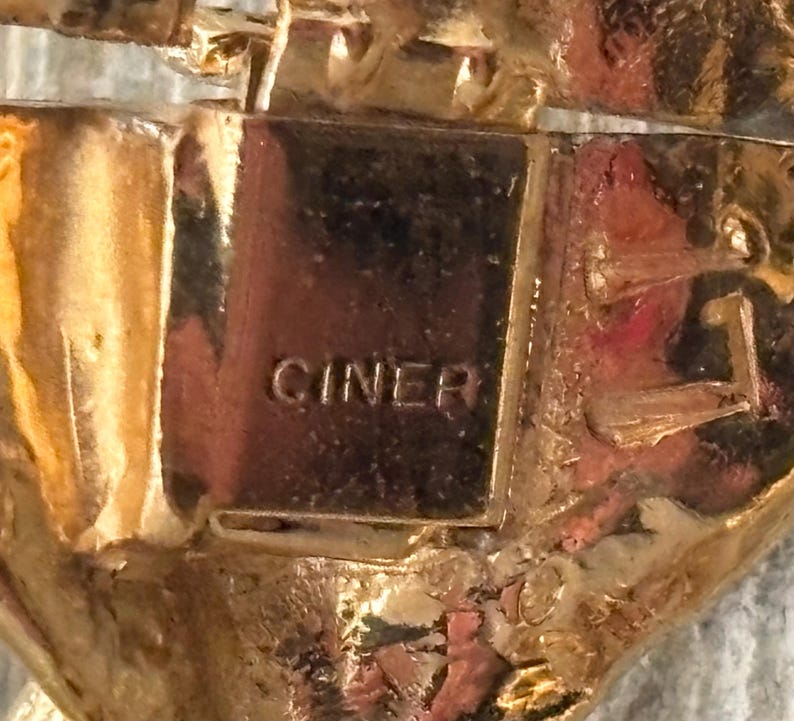 Pu&ograve; includere: Primo piano di un gioiello dorato con la scritta "CINER" stampata su una placca rettangolare. Il gioiello presenta un design ornato e strutturato con dettagli intricati e una finitura lucida.