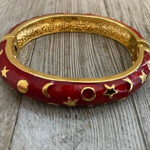 May include: A red enamel bangle bracelet with gold accents. The bracelet is decorated with a pattern of stars, moons, and circles.