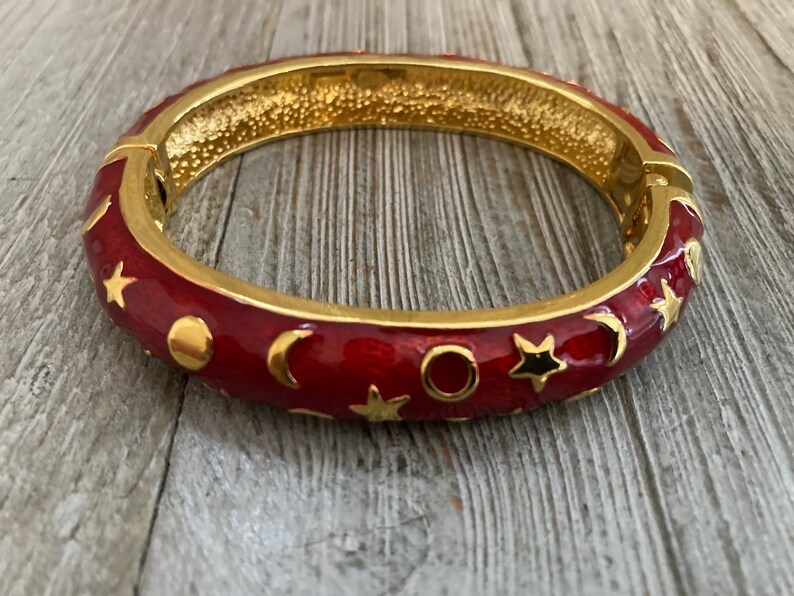 May include: A red enamel bangle bracelet with gold accents. The bracelet is decorated with a pattern of stars, moons, and circles.