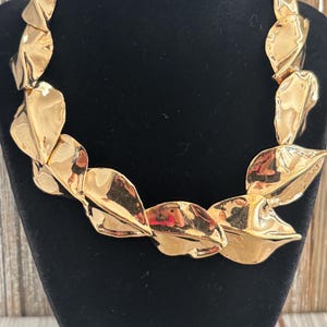 Pu&ograve; includere: Una collana dorata con una serie di pendenti a forma di foglia sovrapposti. La collana &egrave; esposta su un busto di velluto nero, con uno sfondo in legno visibile.