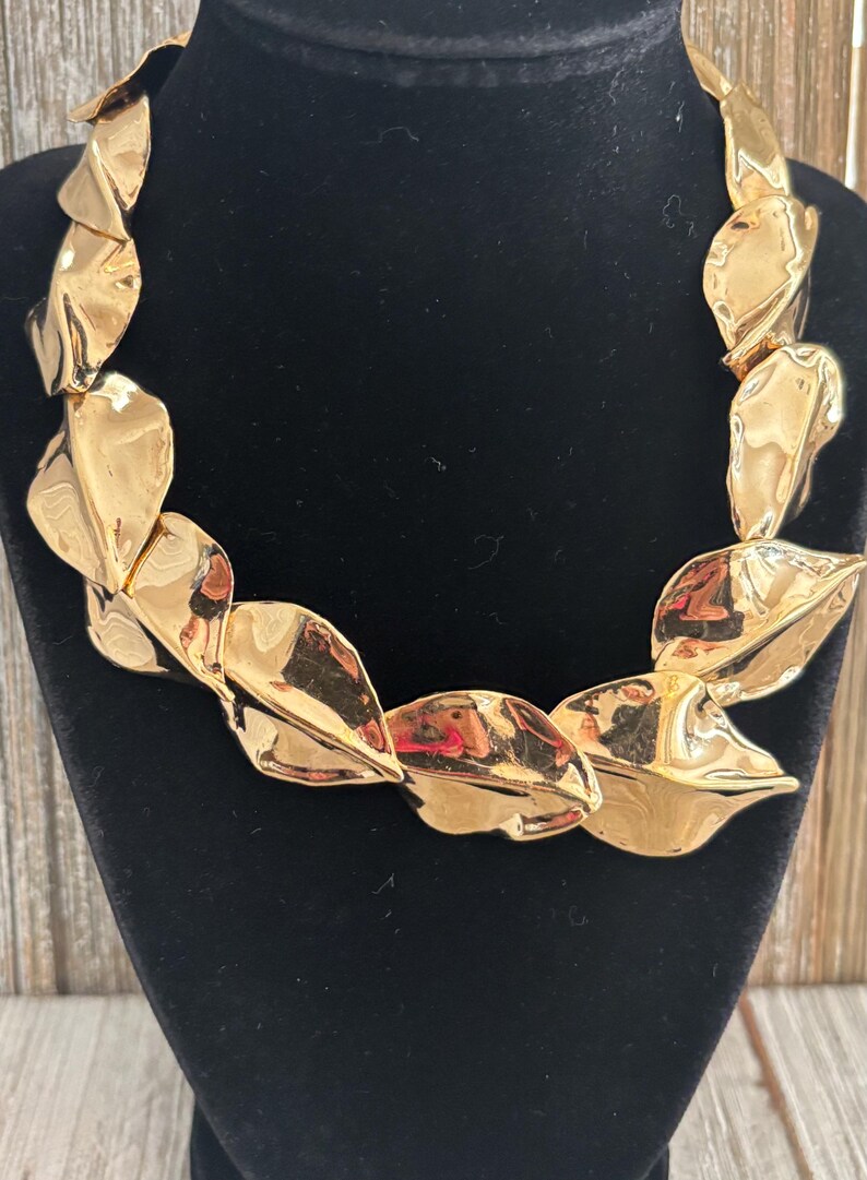 Pu&ograve; includere: Una collana dorata con una serie di pendenti a forma di foglia sovrapposti. La collana &egrave; esposta su un busto di velluto nero, con uno sfondo in legno visibile.