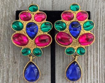 May include: A pair of statement earrings with a gold-tone metal base. The earrings feature a cluster of colorful, faceted gemstones in shades of pink, green, and blue. Each earring has a teardrop-shaped blue gemstone pendant.