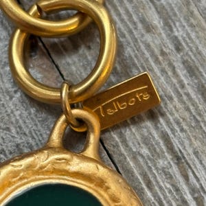 K&ouml;nnte beinhalten: Nahaufnahme eines goldfarbenen Anh&auml;ngers mit gr&uuml;nem Mittelteil und dem Markennamen "Talbots" auf einem kleinen rechteckigen Anh&auml;nger. Der Anh&auml;nger ist an einer goldenen Kette befestigt und zeigt ein Vintage-Schmuckdesign.