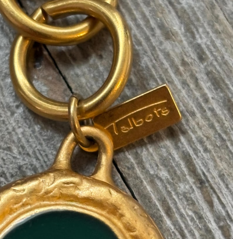 K&ouml;nnte beinhalten: Nahaufnahme eines goldfarbenen Anh&auml;ngers mit gr&uuml;nem Mittelteil und dem Markennamen "Talbots" auf einem kleinen rechteckigen Anh&auml;nger. Der Anh&auml;nger ist an einer goldenen Kette befestigt und zeigt ein Vintage-Schmuckdesign.