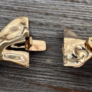 Pu&ograve; includere: Primo piano di una chiusura di collana dorata, slacciata. La chiusura &egrave; realizzata in un materiale metallico lucido, con un design dettagliato. Lo sfondo &egrave; una superficie di legno invecchiato.