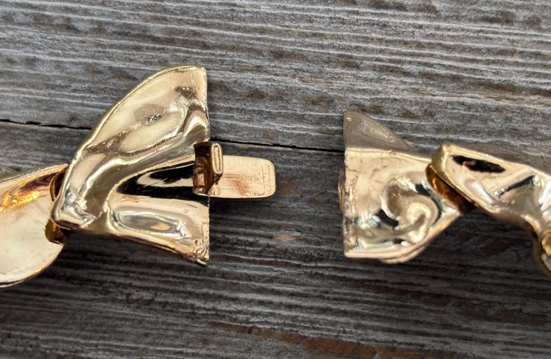 Pu&ograve; includere: Primo piano di una chiusura di collana dorata, slacciata. La chiusura &egrave; realizzata in un materiale metallico lucido, con un design dettagliato. Lo sfondo &egrave; una superficie di legno invecchiato.