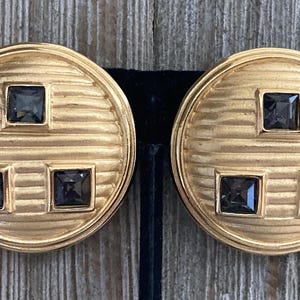 Peut inclure: Une paire de boucles d'oreilles clips dorées à l'aspect géométrique avec des pierres carrées noires.