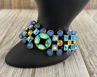 May include: A statement bracelet featuring a mosaic of blue, yellow, and iridescent gemstones. The bracelet is composed of various shapes, including circles and squares, set in a silver-toned metal. The design is geometric and eye-catching.