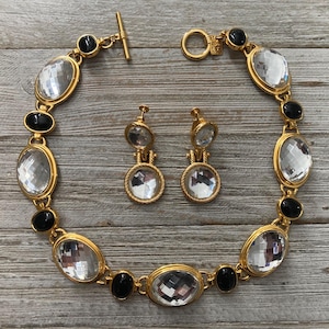Pode incluir: Um colar dourado com pedras ovais pretas e transparentes. Brincos combinando com pedras redondas transparentes.