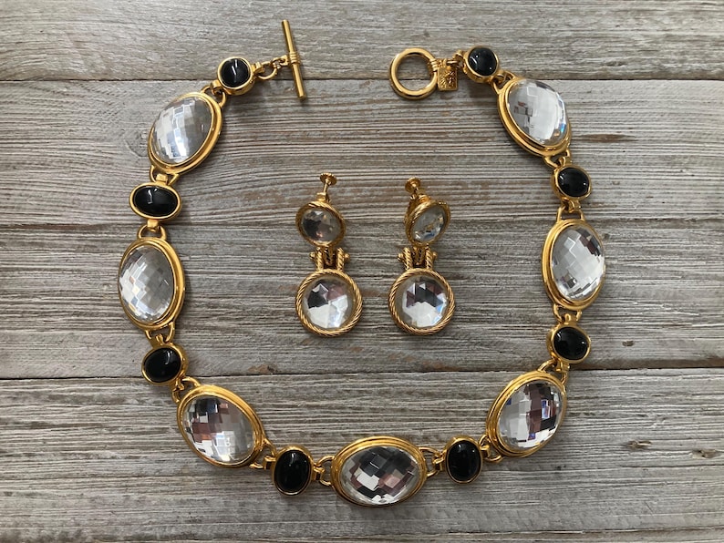 Pode incluir: Um colar dourado com pedras ovais pretas e transparentes. Brincos combinando com pedras redondas transparentes.
