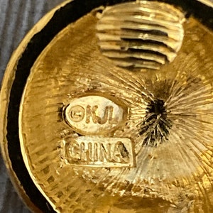 May include: Close-up of a gold-toned circular earring with a black rim. The earring has a textured surface and the words "&copy;KJL CHINA" are stamped on the back.