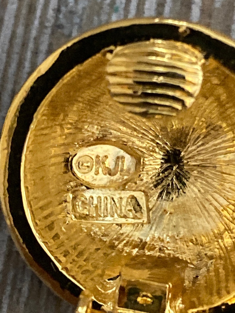 May include: Close-up of a gold-toned circular earring with a black rim. The earring has a textured surface and the words "&copy;KJL CHINA" are stamped on the back.