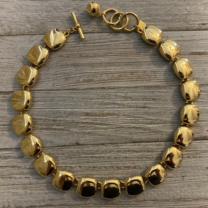 May include: A gold-toned necklace featuring large, square-shaped beads. The necklace has a toggle clasp and a series of small rings connecting the chain to the clasp. The beads have a textured surface and reflect light, creating a shiny appearance.