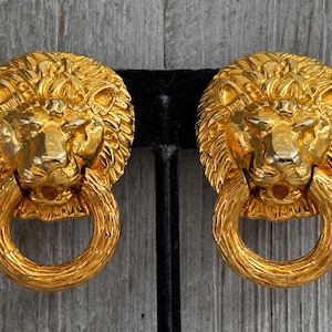 Puede incluir: Un par de pendientes dorados con cabeza de león. Cada pendiente presenta una cara de león detallada con un elemento en forma de anillo colgando debajo. Los pendientes se exhiben en un soporte negro sobre un fondo de madera.
