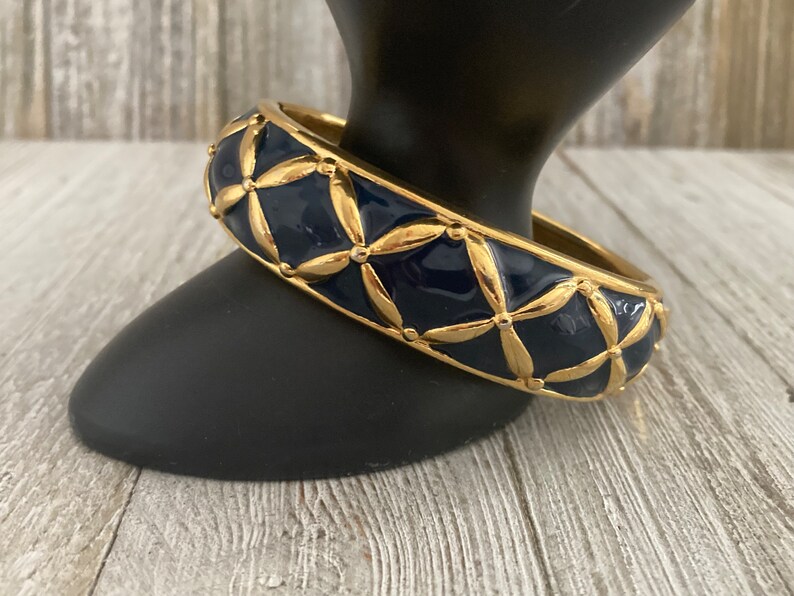 May include: A gold bangle bracelet with a navy blue enamel finish. The bracelet features a geometric pattern of gold leaves and dots.