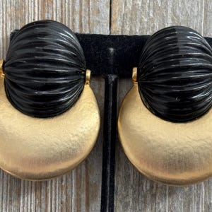 May include: A pair of statement earrings. Each earring features a half-circle of brushed gold-tone metal topped with a black, ridged, oval-shaped element. The earrings are attached with small gold-tone connectors.