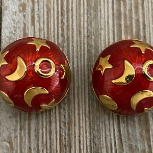 May include: A pair of red and gold earrings with a celestial design. The earrings are round and feature a pattern of stars and crescent moons.