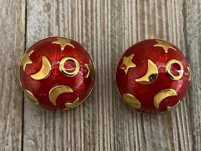 May include: A pair of red and gold earrings with a celestial design. The earrings are round and feature a pattern of stars and crescent moons.
