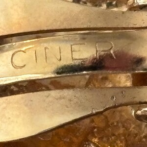 Pu&ograve; includere: Primo piano di una chiusura di gioiello dorata con la scritta incisa "CINER". La chiusura ha un design curvo con segni di usura visibili, che suggeriscono che sia vintage. Lo sfondo &egrave; sfocato, concentrando l'attenzione sulla chiusura.