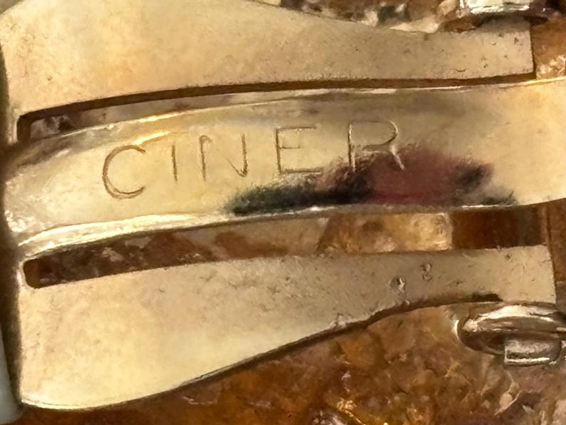 Pu&ograve; includere: Primo piano di una chiusura di gioiello dorata con la scritta incisa "CINER". La chiusura ha un design curvo con segni di usura visibili, che suggeriscono che sia vintage. Lo sfondo &egrave; sfocato, concentrando l'attenzione sulla chiusura.