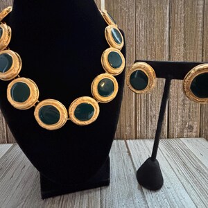 K&ouml;nnte beinhalten: Ein goldfarbenes Halsketten- und Ohrring-Set. Die Halskette und die Ohrringe haben gro&szlig;e, kreisf&ouml;rmige, dunkelgr&uuml;ne Emaille-Mittelst&uuml;cke, die von einem strukturierten goldfarbenen Metall umgeben sind. Der Schmuck wird auf einer schwarzen Samtb&uuml;ste und einem Ohrringst&auml;nder pr&auml;sentiert.