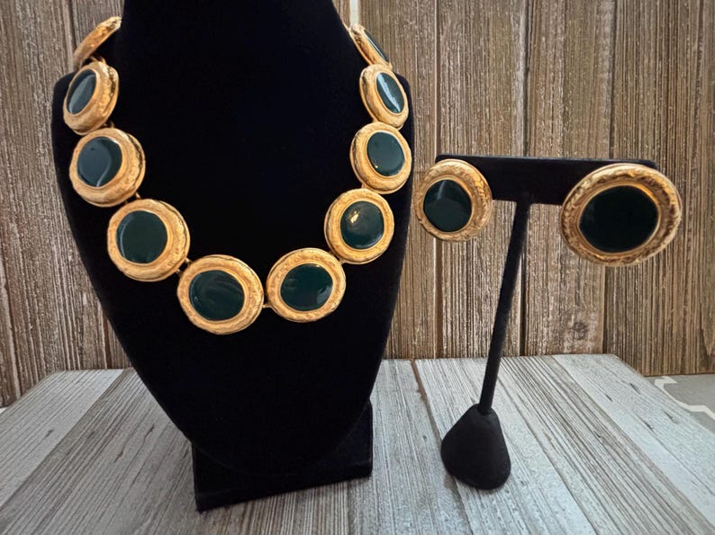 K&ouml;nnte beinhalten: Ein goldfarbenes Halsketten- und Ohrring-Set. Die Halskette und die Ohrringe haben gro&szlig;e, kreisf&ouml;rmige, dunkelgr&uuml;ne Emaille-Mittelst&uuml;cke, die von einem strukturierten goldfarbenen Metall umgeben sind. Der Schmuck wird auf einer schwarzen Samtb&uuml;ste und einem Ohrringst&auml;nder pr&auml;sentiert.