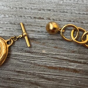 K&ouml;nnte beinhalten: Nahaufnahme einer goldfarbenen Halskette mit ovalen, dunkelgr&uuml;nen Edelsteinakzenten. Die Halskette verf&uuml;gt &uuml;ber einen Knebelverschluss und eine Reihe ineinandergreifender Ringe. Der Schmuck liegt auf einer verwitterten Holzoberfl&auml;che.