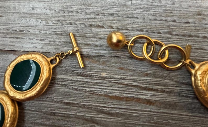 K&ouml;nnte beinhalten: Nahaufnahme einer goldfarbenen Halskette mit ovalen, dunkelgr&uuml;nen Edelsteinakzenten. Die Halskette verf&uuml;gt &uuml;ber einen Knebelverschluss und eine Reihe ineinandergreifender Ringe. Der Schmuck liegt auf einer verwitterten Holzoberfl&auml;che.