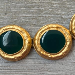 K&ouml;nnte beinhalten: Nahaufnahme einer goldfarbenen Halskette mit kreisf&ouml;rmigen Gliedern. Jedes Glied hat eine dunkelgr&uuml;ne Mitte und einen strukturierten, verzierten Goldrand. Die Halskette wird auf einer hellgrauen Holzoberfl&auml;che pr&auml;sentiert.