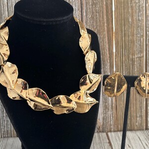Pu&ograve; includere: Un set di collana e orecchini color oro. La collana e gli orecchini presentano un design a foglie con una superficie strutturata. I gioielli sono esposti su un manichino nero e un supporto, su sfondo di legno.