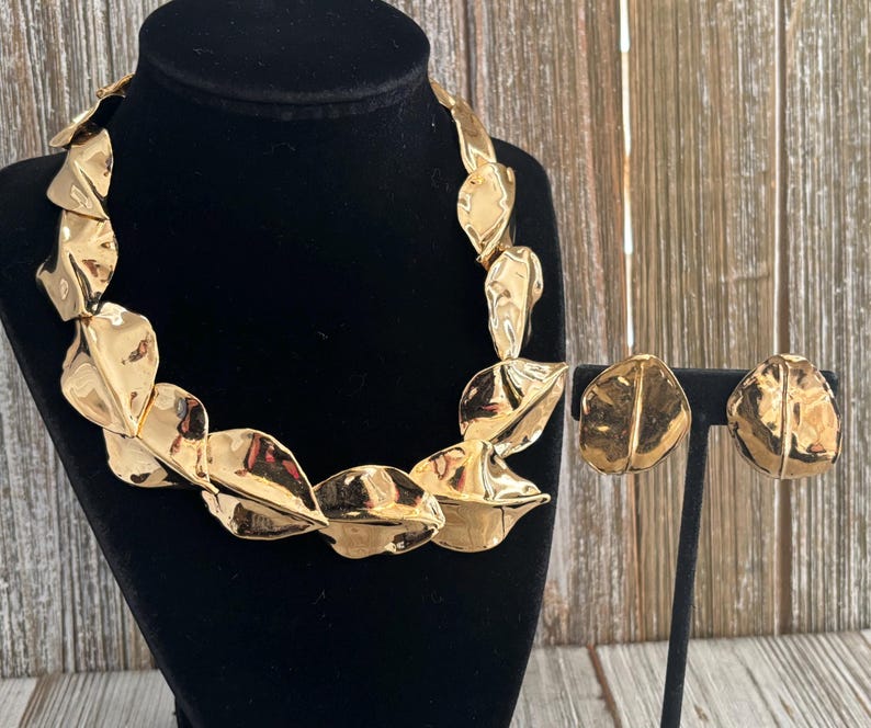 Pu&ograve; includere: Un set di collana e orecchini color oro. La collana e gli orecchini presentano un design a foglie con una superficie strutturata. I gioielli sono esposti su un manichino nero e un supporto, su sfondo di legno.