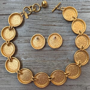K&ouml;nnte beinhalten: Goldfarbene Halskette und passende Ohrstecker. Die Halskette besteht aus einer Reihe von verbundenen kreisf&ouml;rmigen Medaillons. Die Ohrringe sind einfache, runde Stecker. Der Schmuck wird auf einer verwitterten, grauen Holzoberfl&auml;che pr&auml;sentiert.