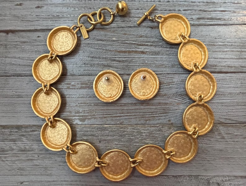 K&ouml;nnte beinhalten: Goldfarbene Halskette und passende Ohrstecker. Die Halskette besteht aus einer Reihe von verbundenen kreisf&ouml;rmigen Medaillons. Die Ohrringe sind einfache, runde Stecker. Der Schmuck wird auf einer verwitterten, grauen Holzoberfl&auml;che pr&auml;sentiert.