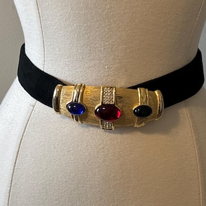 Op de afbeelding: Een zwarte riem met een brede, getextureerde gesp van goudkleurig metaal. De gesp is versierd met drie ovale cabochonstenen in blauw, rood en zwart, geaccentueerd door kleine heldere kristallen in verticale rijen tussen de stenen.