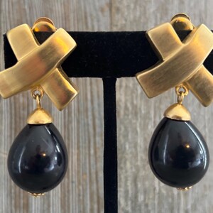 May include: A pair of clip-on earrings featuring a brushed gold-tone metal X-shaped top piece. A smooth, glossy black teardrop-shaped bead hangs from each gold-tone component, creating a dangling design with a polished finish and gold-tone hardware.