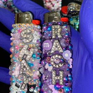 Könnte beinhalten: Zwei personalisierte Feuerzeuge mit Strasssteinen und Charms. Das Feuerzeug links sagt "SHAKY" und hat ein rosa, blaues und weißes Farbschema. Das Feuerzeug rechts sagt "RACH" und hat ein lila, blaues und weißes Farbschema.