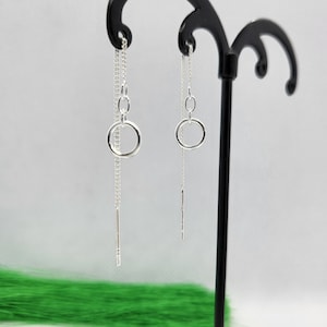 May include: A pair of silver dangle earrings with a small circle charm and a thin chain hanging from each. The earrings are displayed on a black earring stand.
