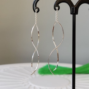 Könnte beinhalten: Ein Paar silberne Ohrringe mit einem gedrehten, unendlichkeitsförmigen Design. Die Ohrringe hängen an einem schwarzen Ständer.