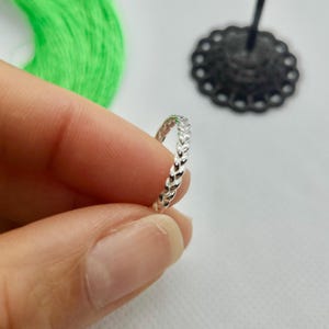 May include: A delicate silver ring with a repeating leaf pattern. The ring is held between fingers, showcasing its intricate design. The ring's polished surface reflects light, highlighting the detailed texture of the leaves.